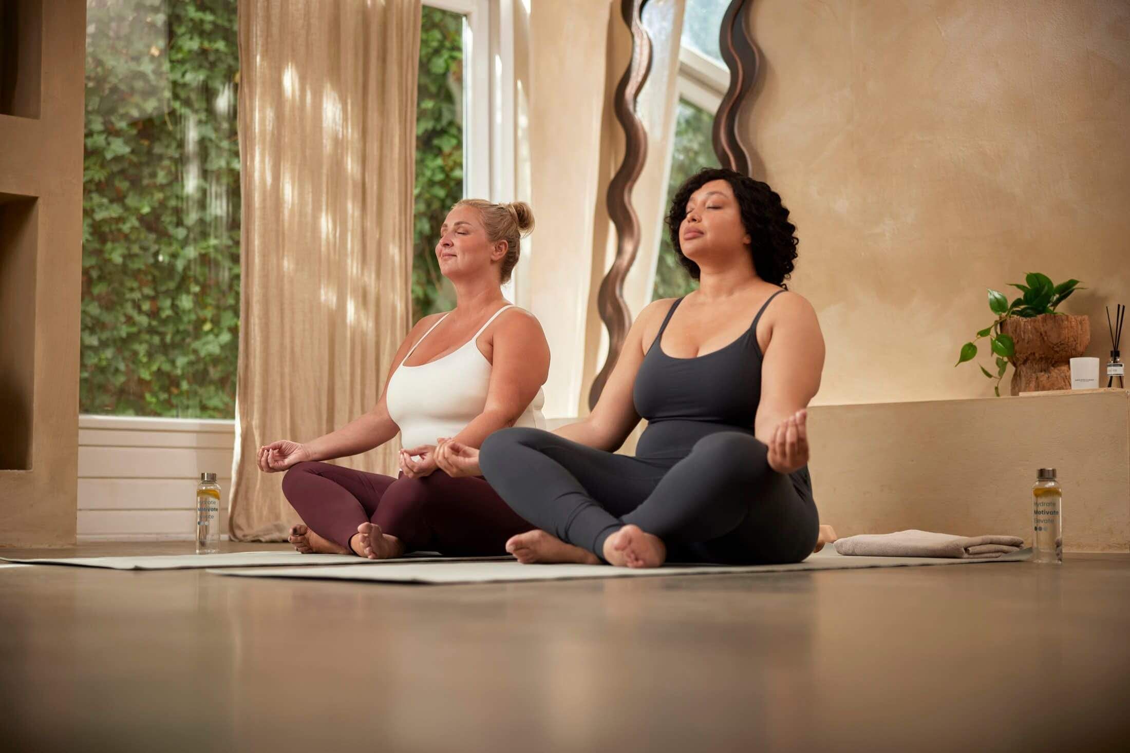 Two women meditate on yoga mats in a sunlit room, eyes closed in peace.