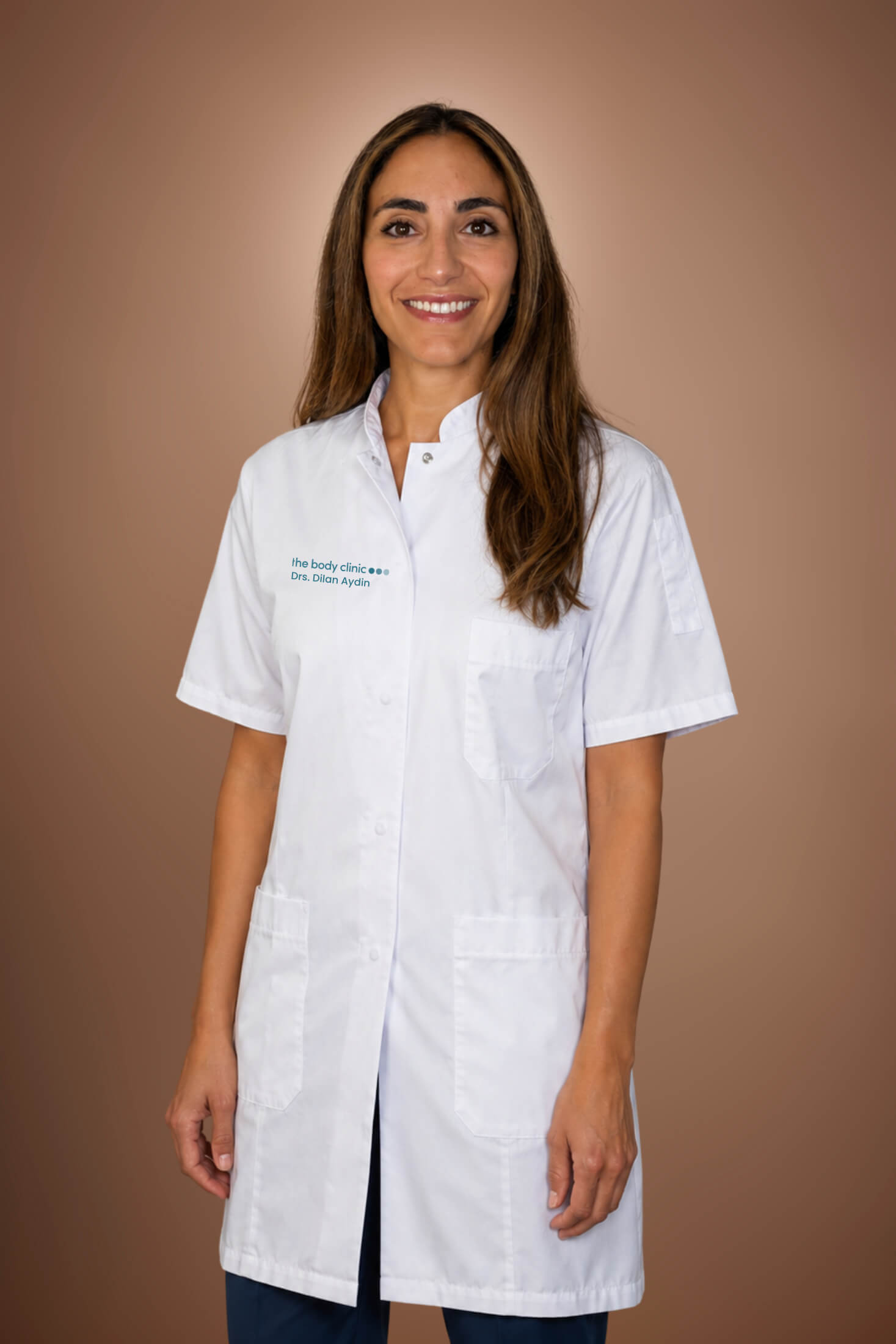 Smiling woman with long dark hair wearing a white medical coat and jeans.
