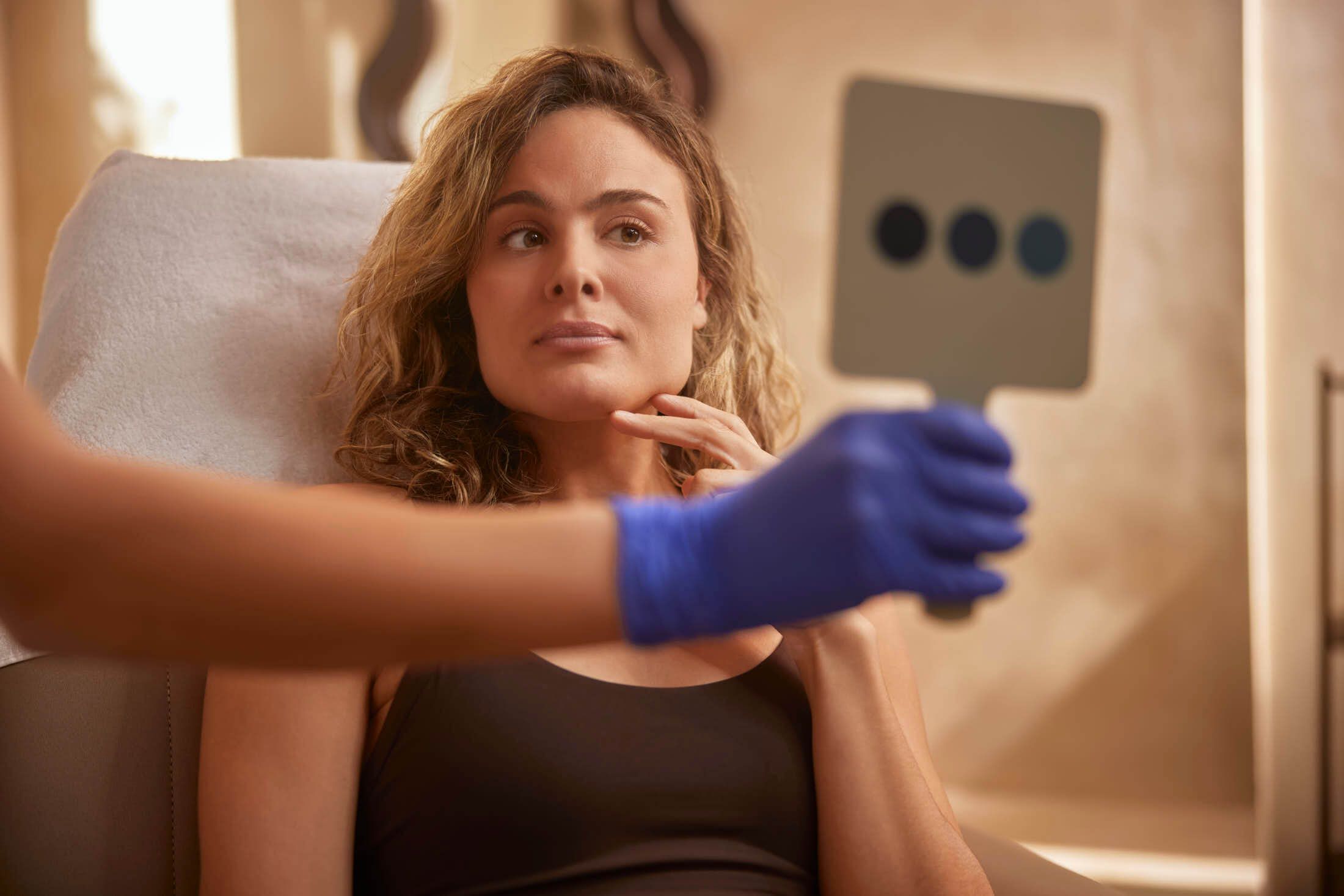 Vrouwelijke client bekijkt zichzelf in de spiegel tijdens voorbereiding op cosmetische behandeling bij The Body Clinic