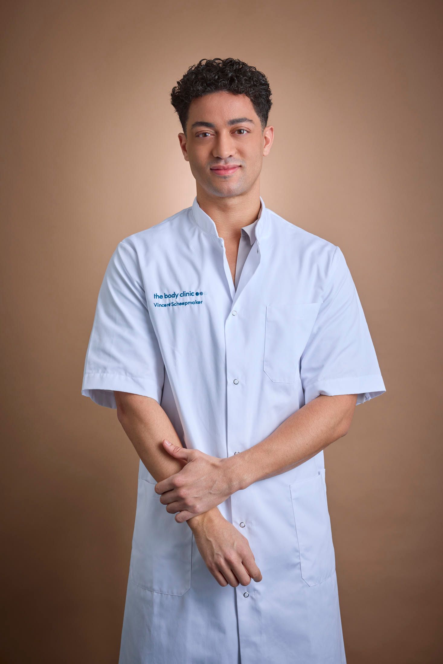 Young man in light blue medical coat stands confidently, looking at the camera.