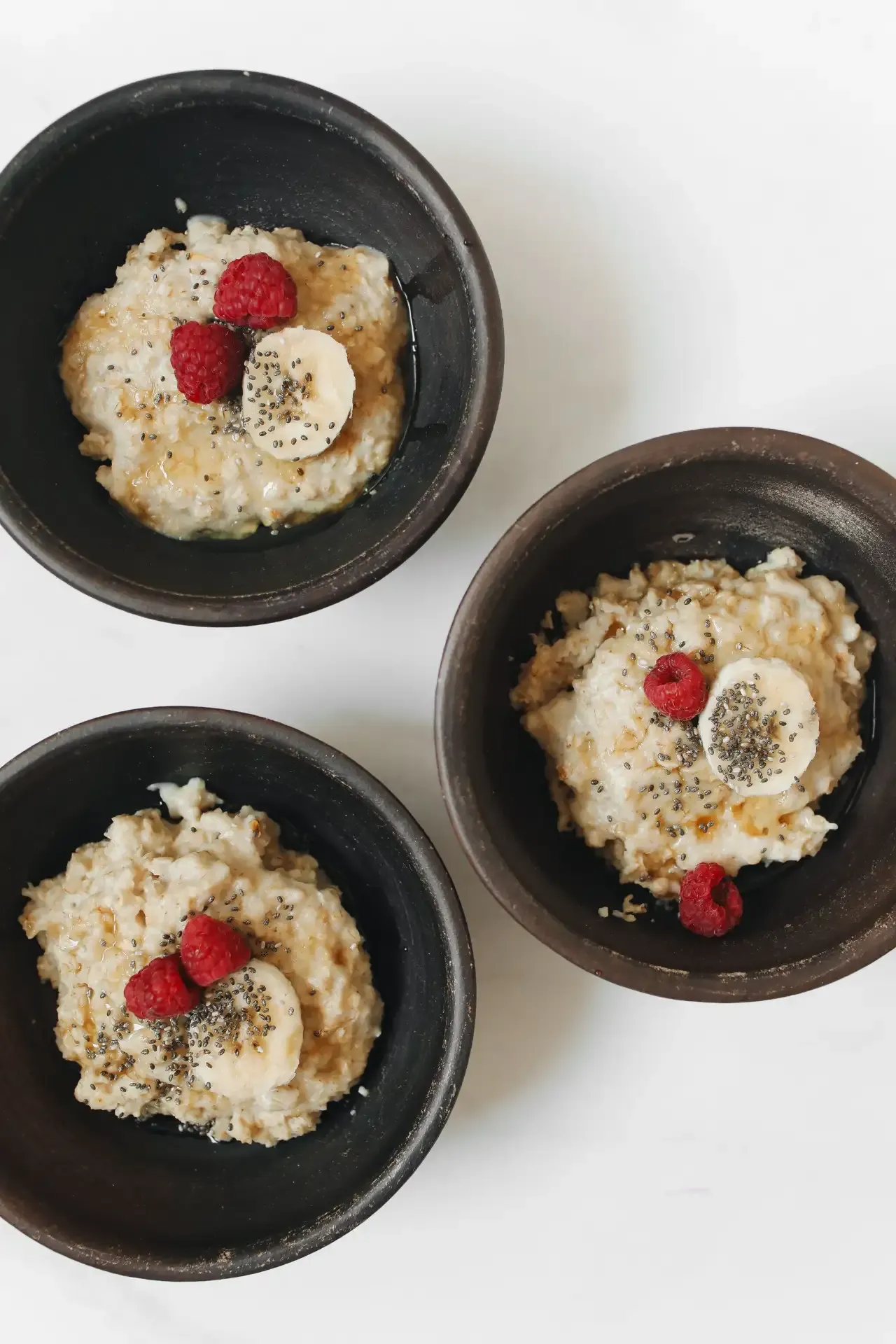 Three small bowls of oatmeal topped with raspberries, banana slices, and chia seeds.