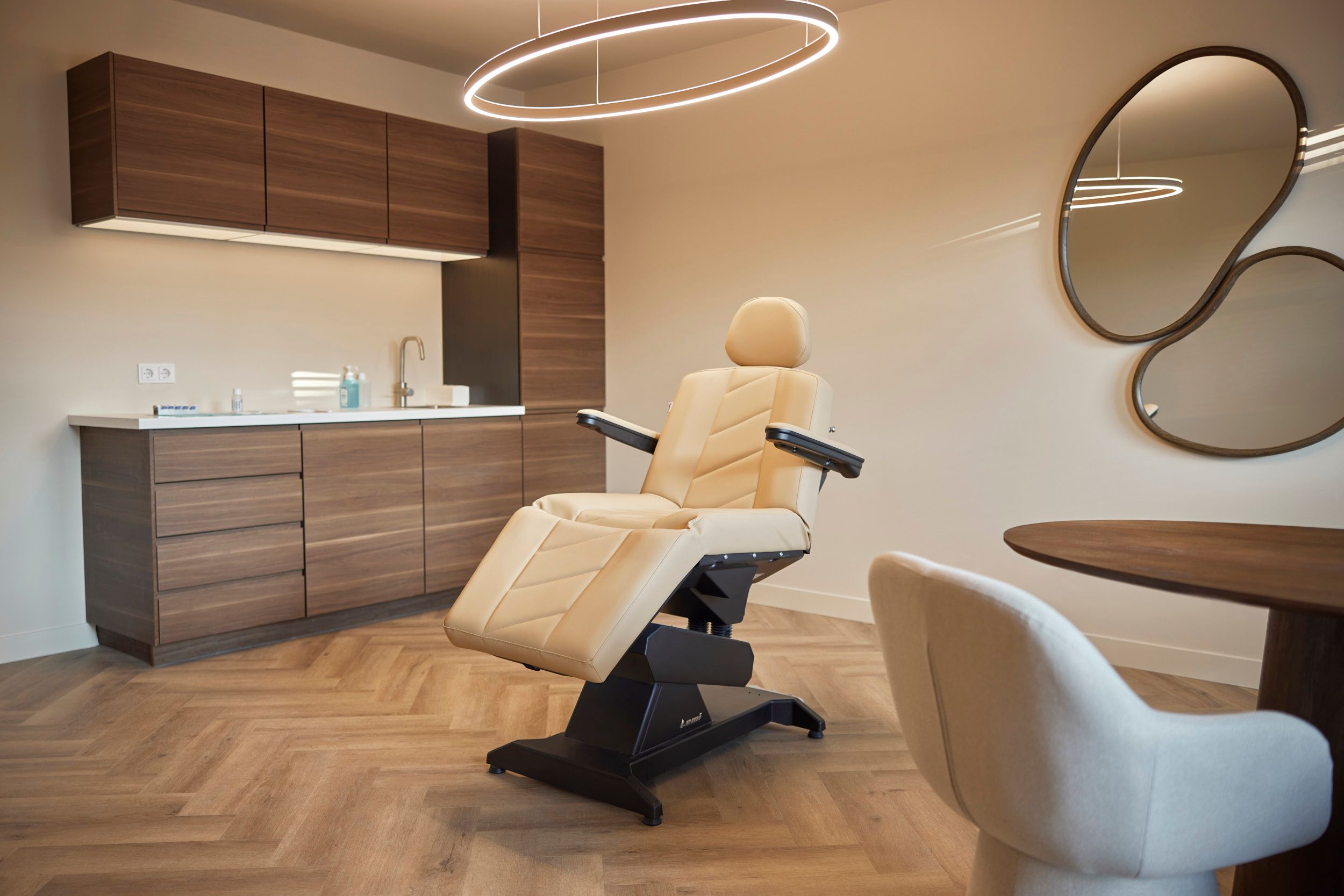 Modern treatment room with beige adjustable chair, wooden cabinets, sink, and abstract mirrors.