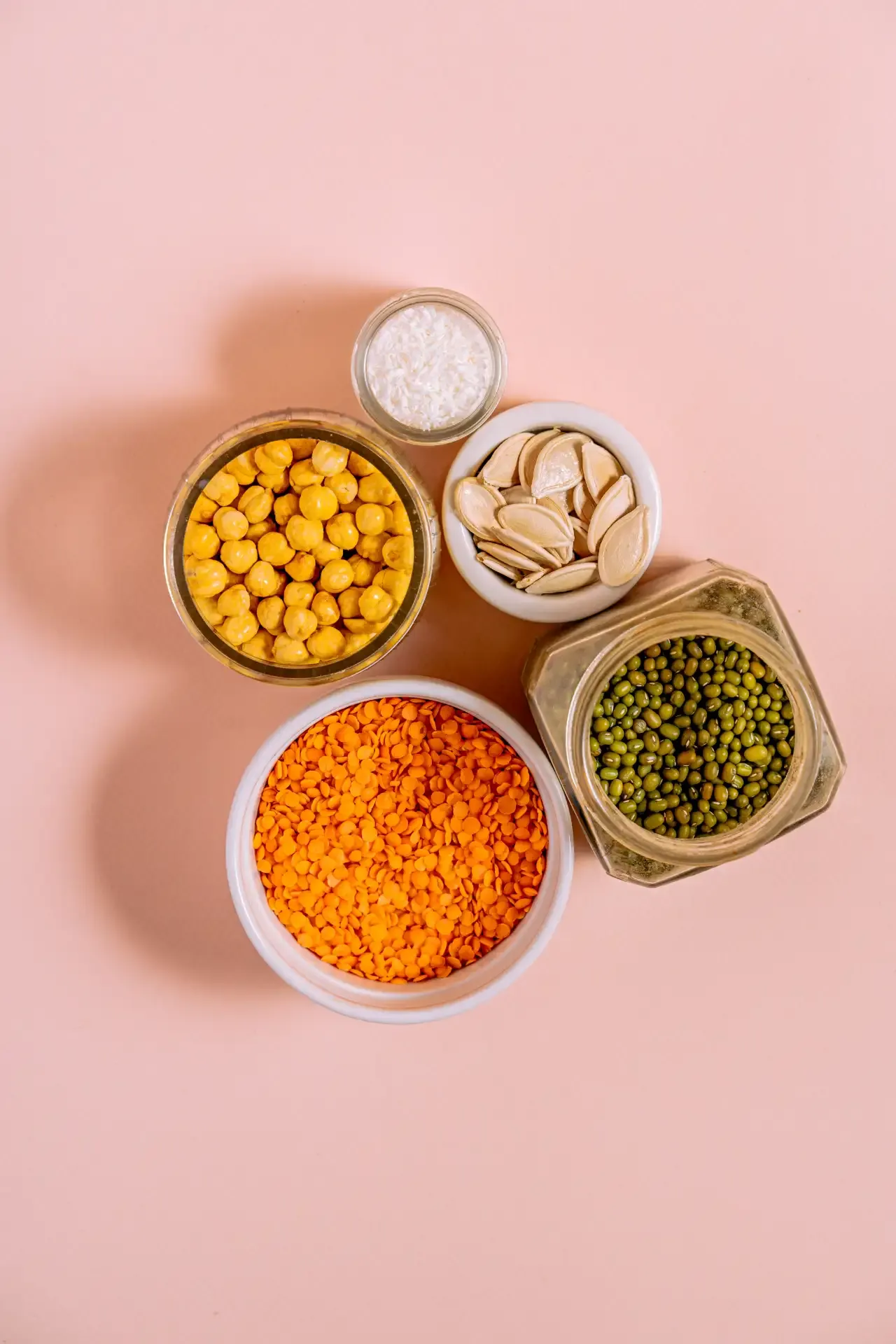 Five bowls containing chickpeas, lentils, mung beans, almonds, and salt on a pink background.