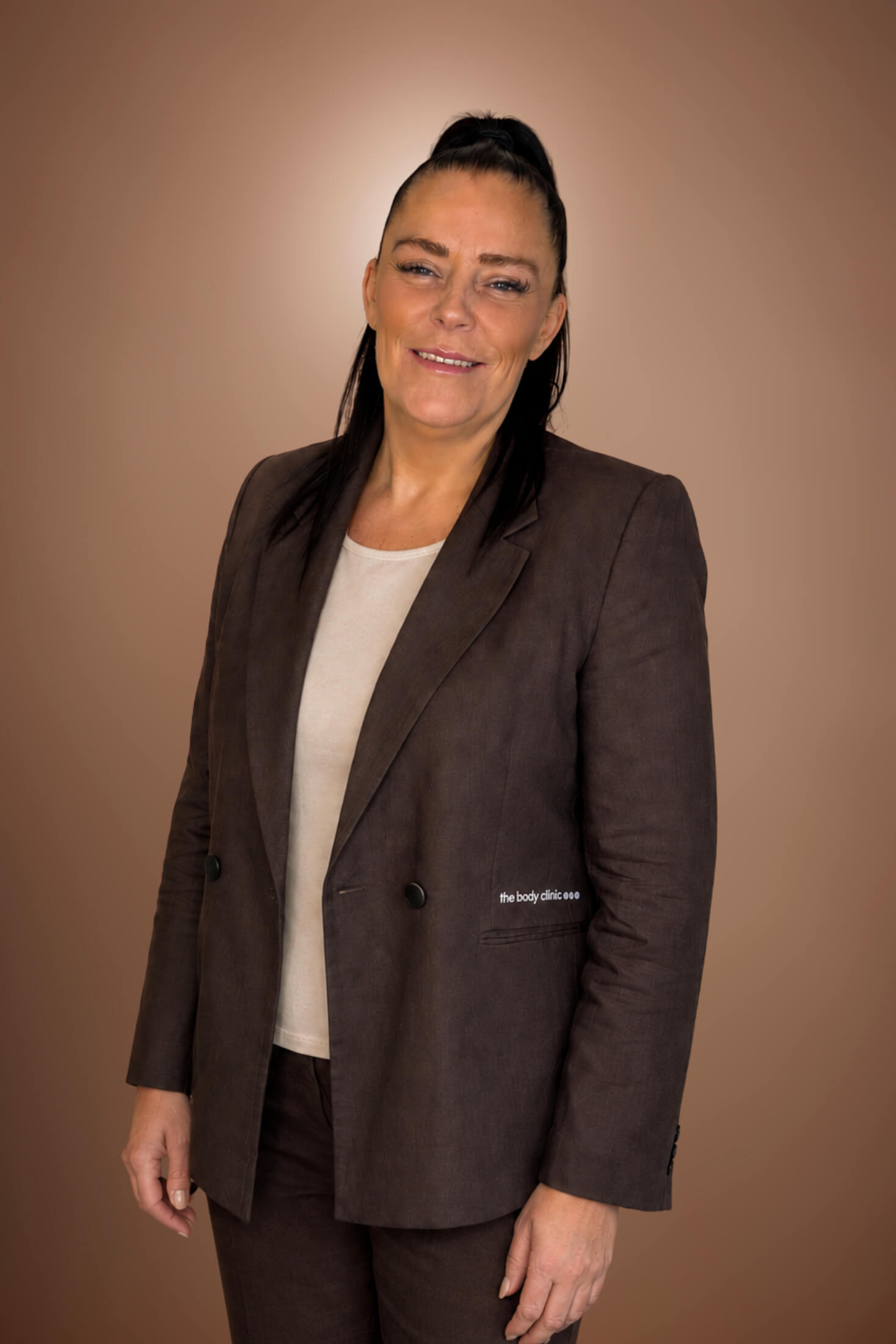 Professional woman with dark hair and a blazer, smiling confidently against a brown background.