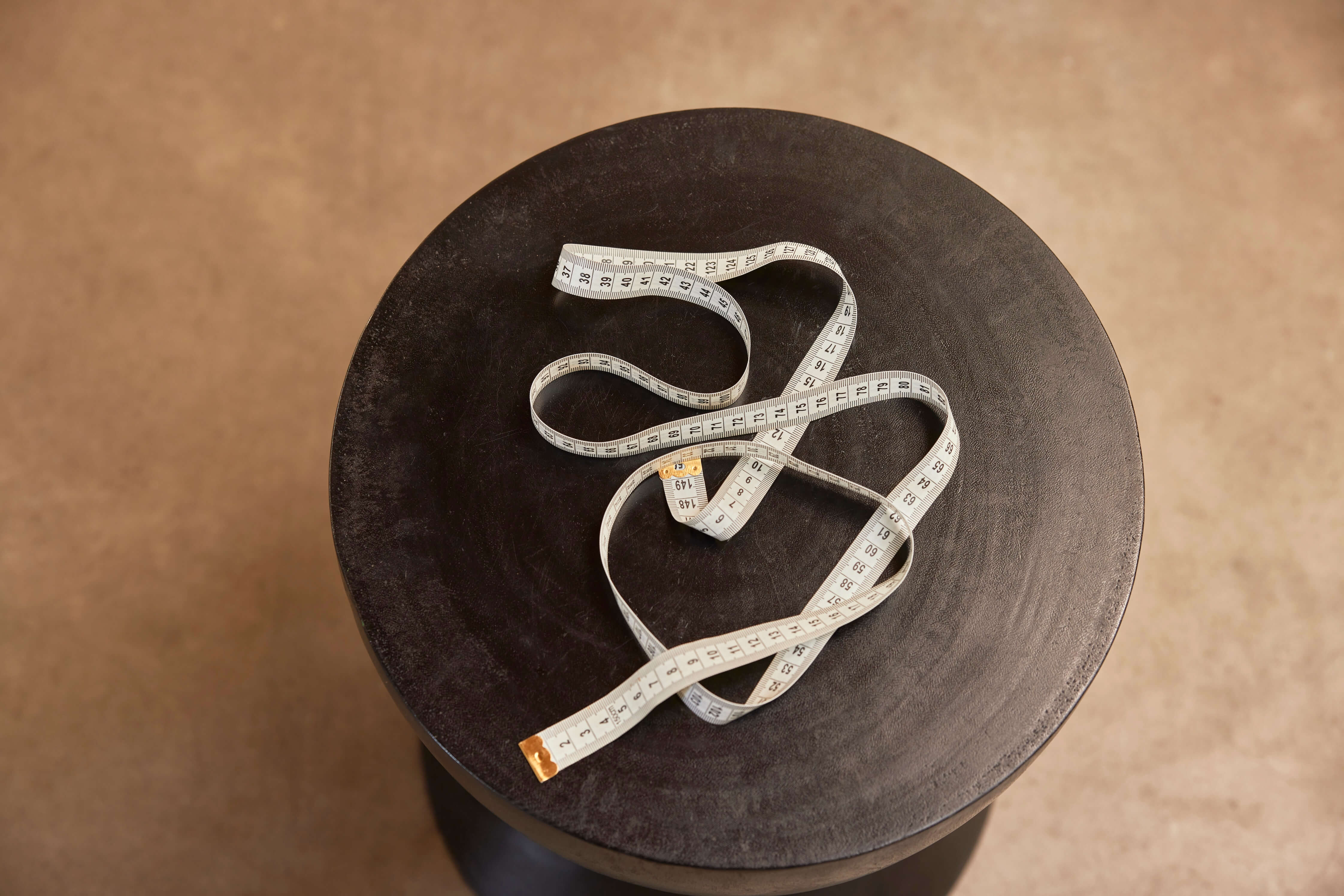 White cloth measuring tape coiled on a dark, round wooden stool top.
