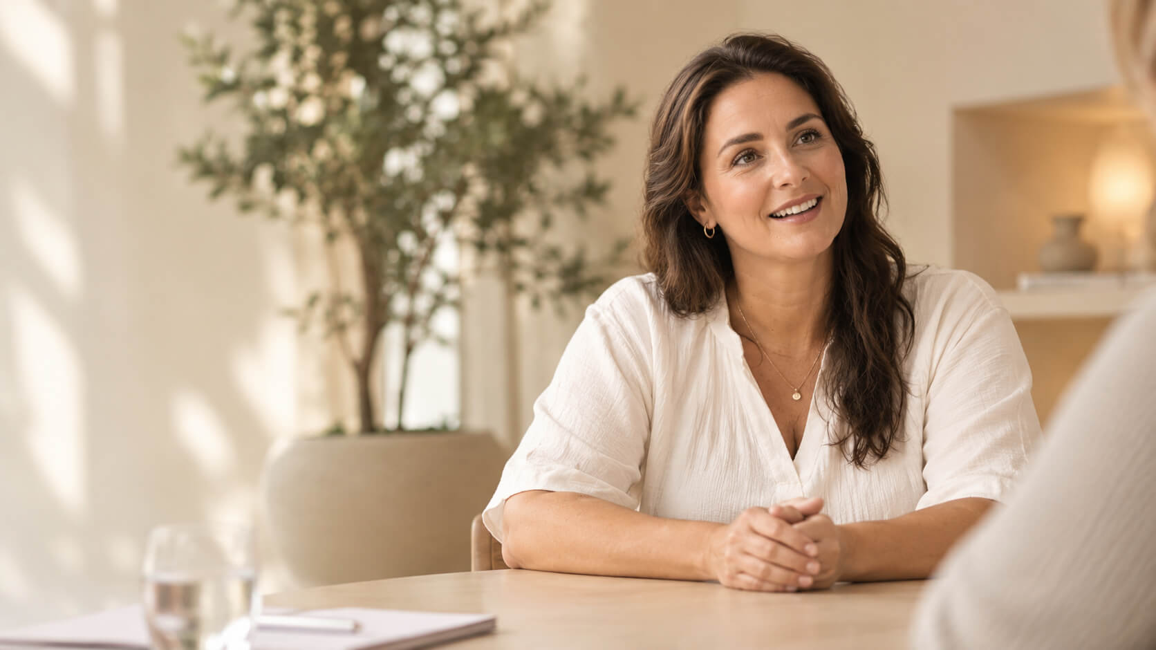 Smiling woman in white shirt talks at a table with clasped hands.