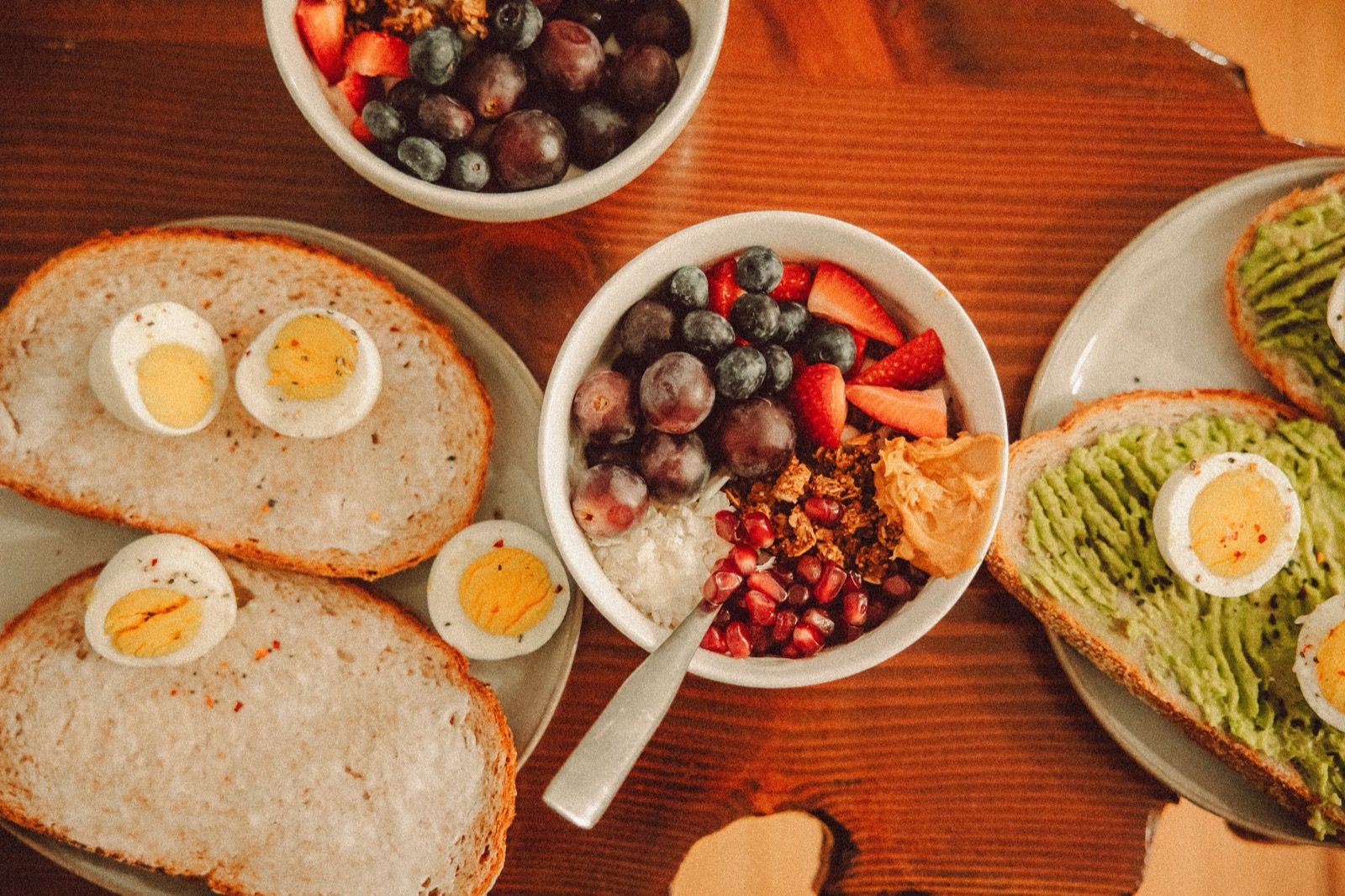 Breakfast spread with boiled eggs, avocado toast, and fruit bowls on a wooden table.