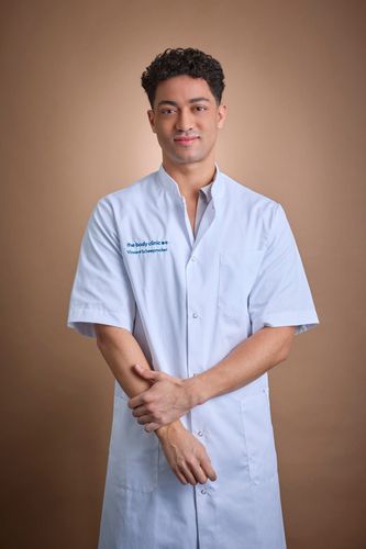 Young man in light blue medical coat stands confidently, looking at the camera.