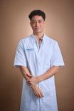 Young man in light blue medical coat stands confidently, looking at the camera.