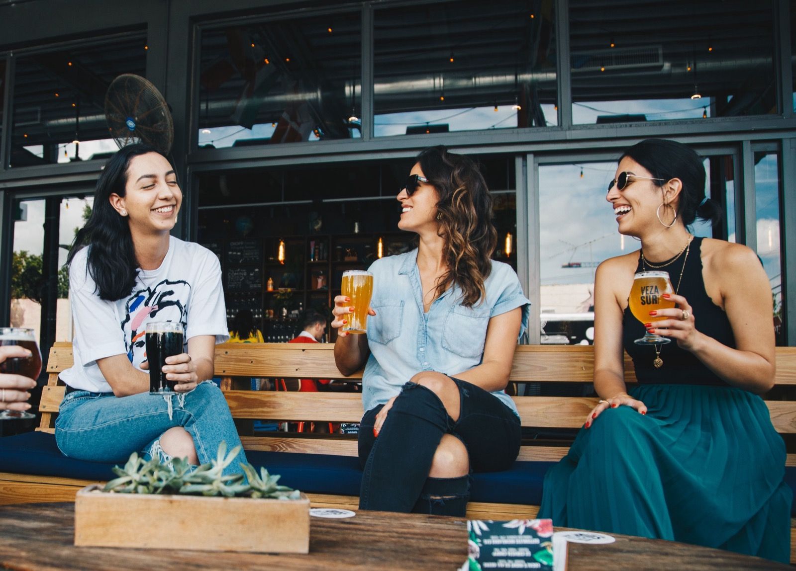 Three women smiling and chatting on an outdoor patio, enjoying drinks.