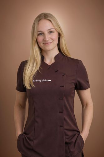 Blonde woman in a dark uniform top, smiling confidently with hands in pockets.