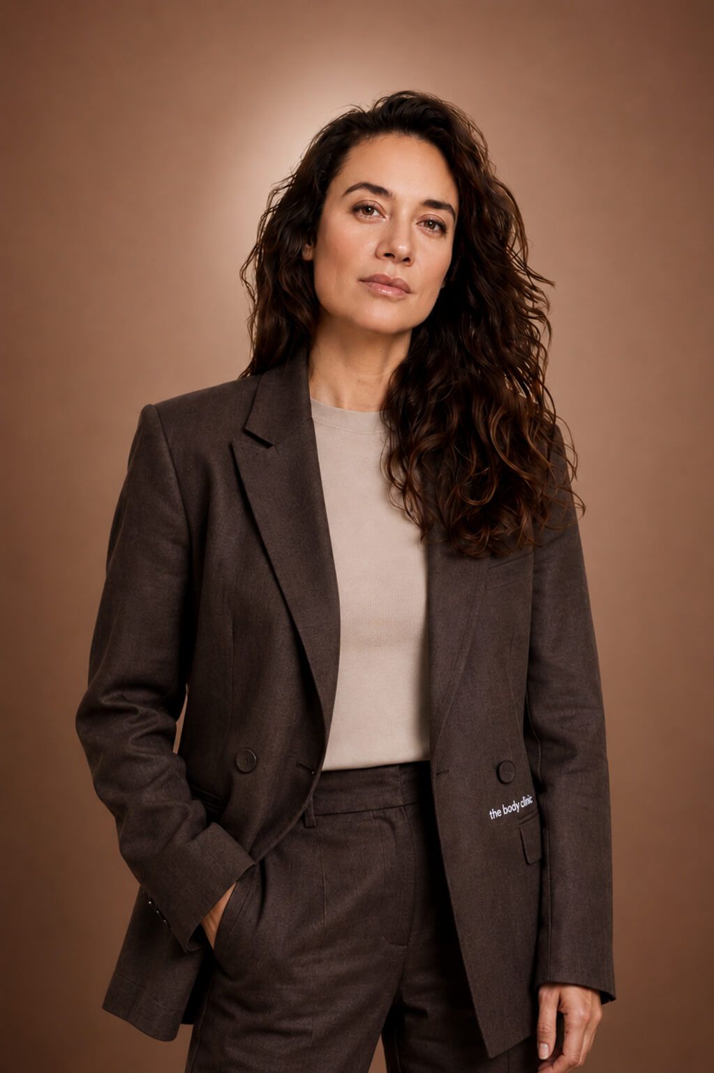 A professional woman with long wavy hair wears a dark brown suit, looking directly ahead.