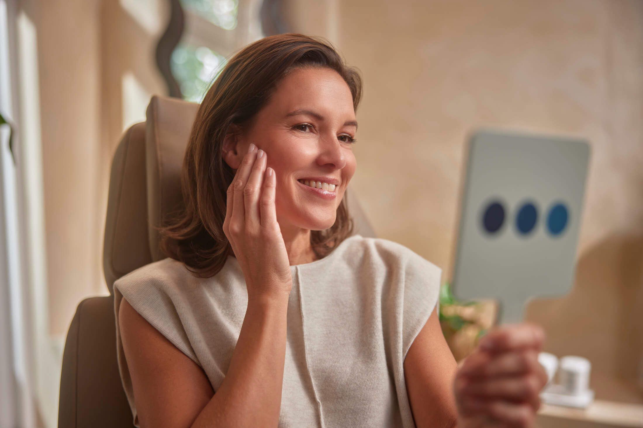 A smiling woman touches her cheek while looking at her reflection in a small mirror.
