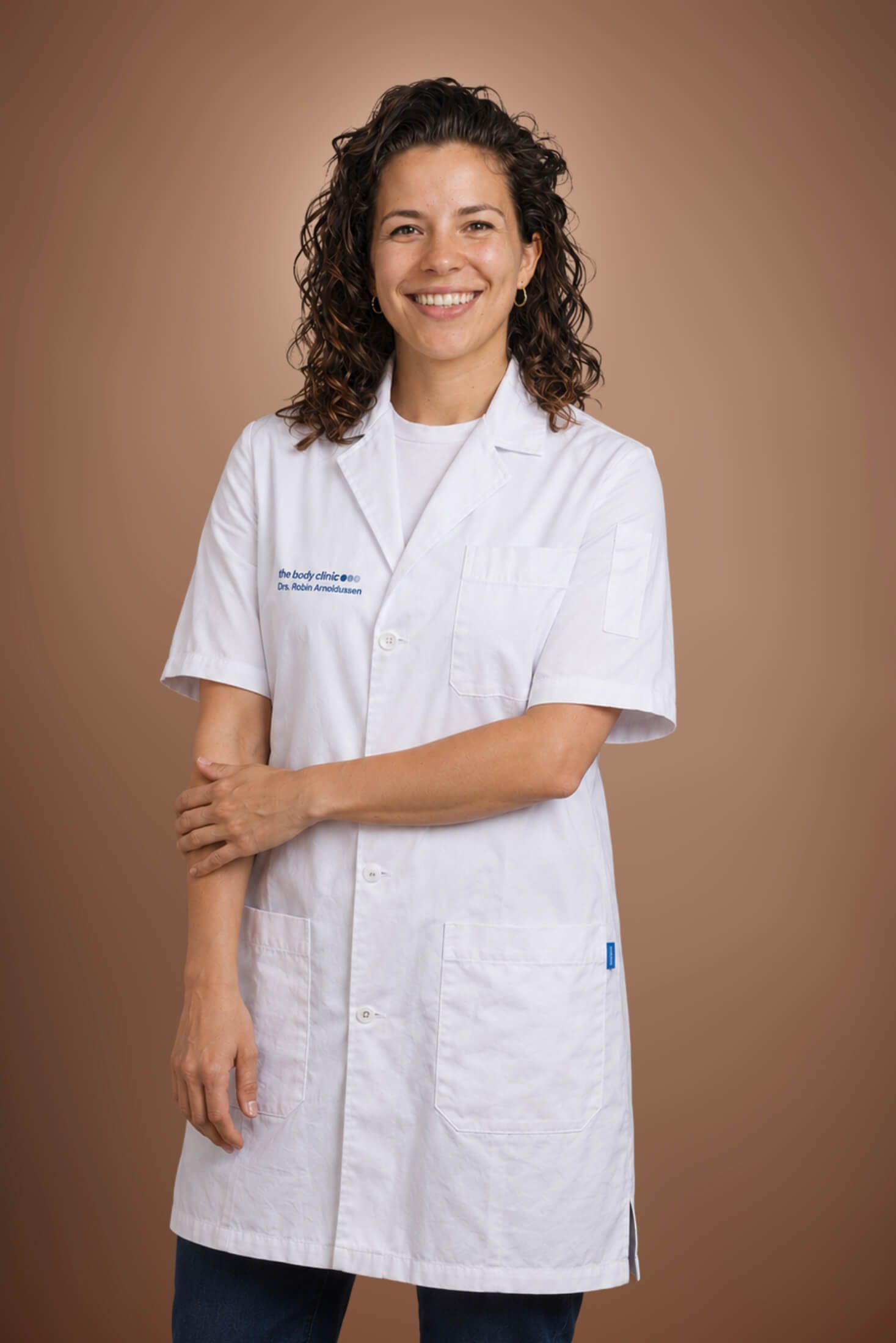 Smiling woman with curly hair wearing a white lab coat against a brown background.