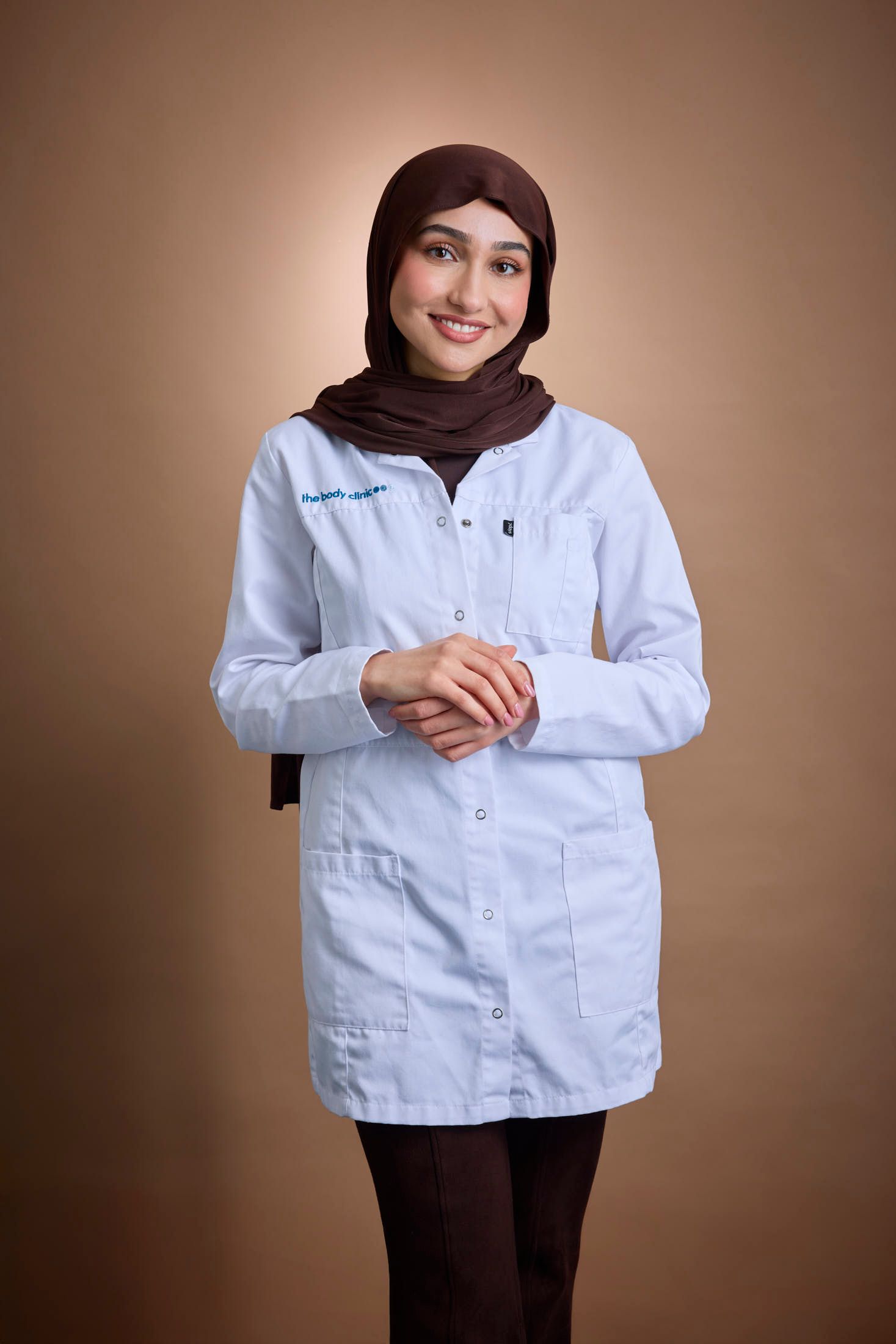 Smiling woman in light blue lab coat and brown hijab with clasped hands.