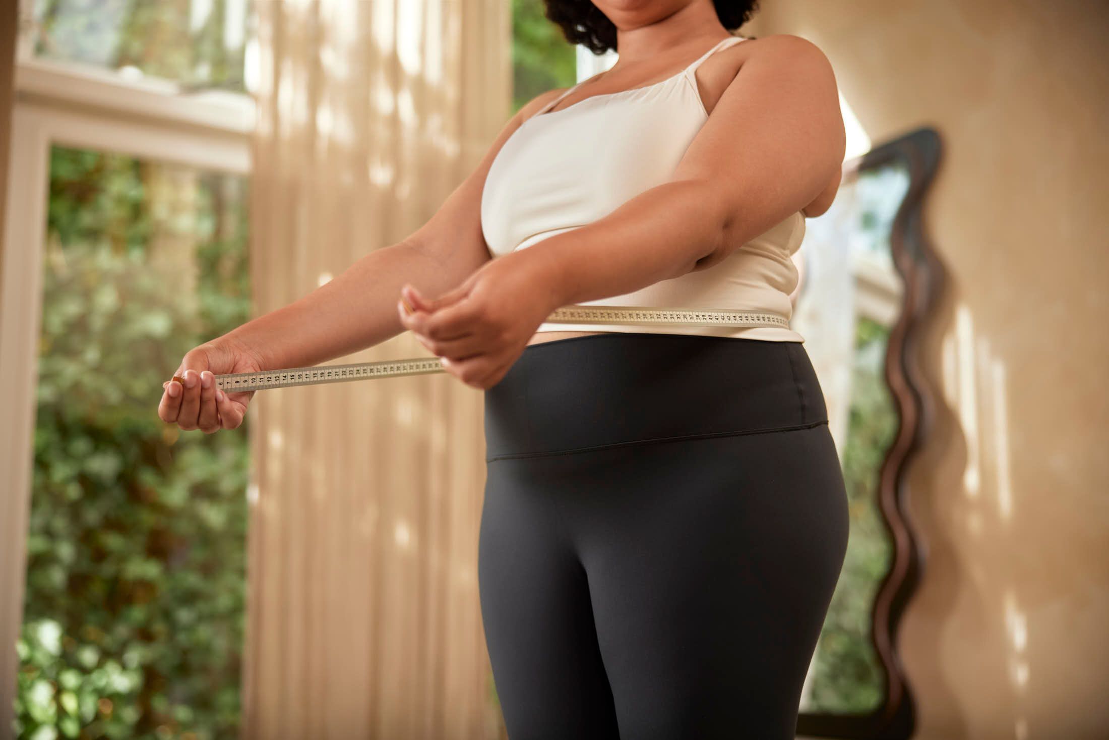 Woman in a white top measuring her waist with a tape measure at home.