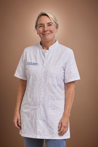 Blonde woman in white medical uniform smiles, standing against a warm brown background.