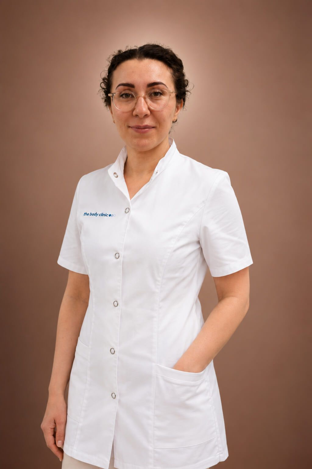 A professional woman in a white medical coat smiles subtly, hands in pockets.