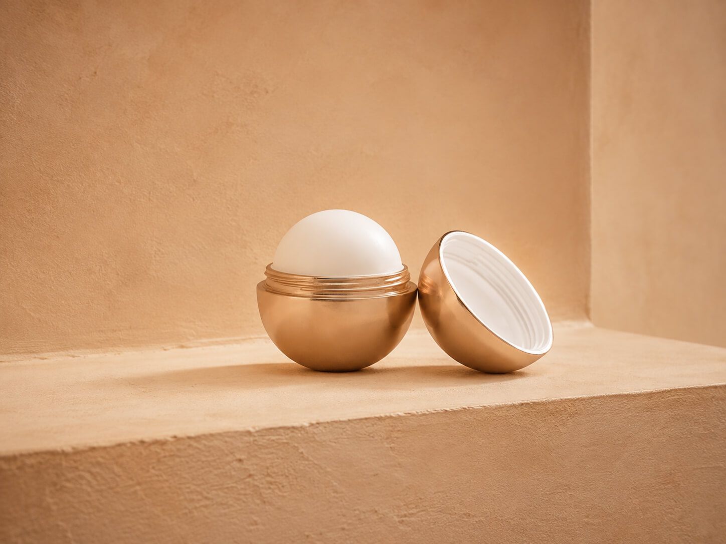 Spherical gold metallic lip balm container open, revealing white product on a warm background.