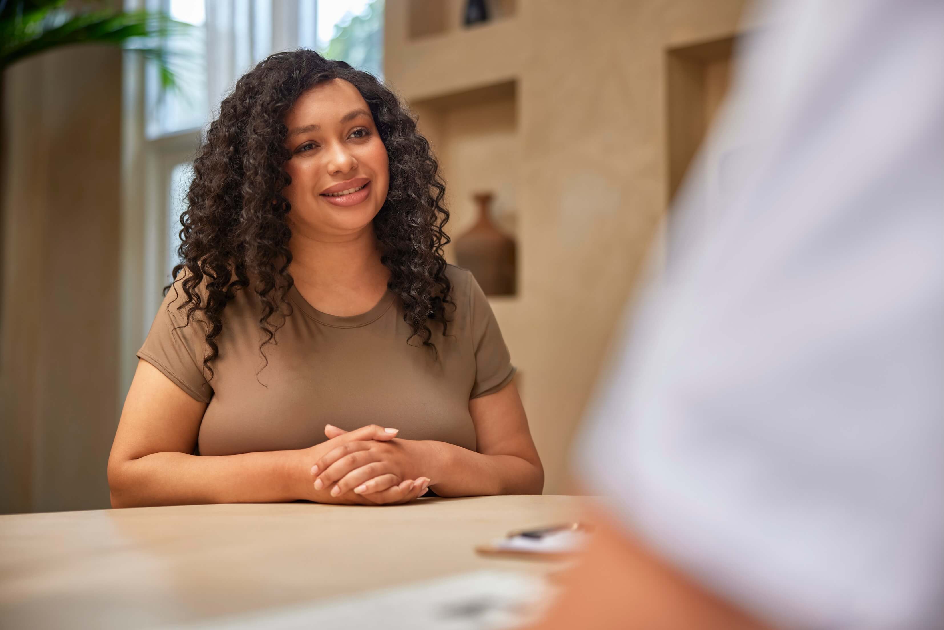A smiling woman with curly dark hair sits at a table, engaging in conversation.