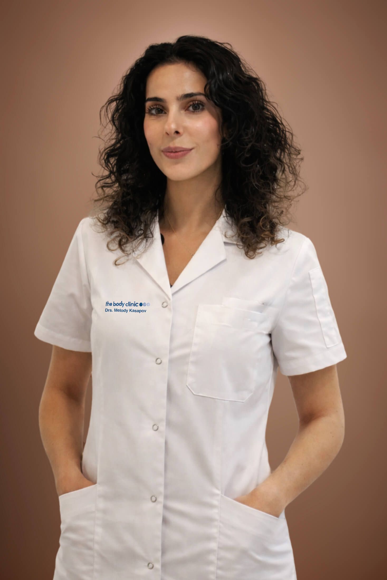 A professional woman with dark curly hair wears a white medical coat, hands in pockets.