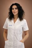 A professional woman with dark curly hair wears a white medical coat, hands in pockets.