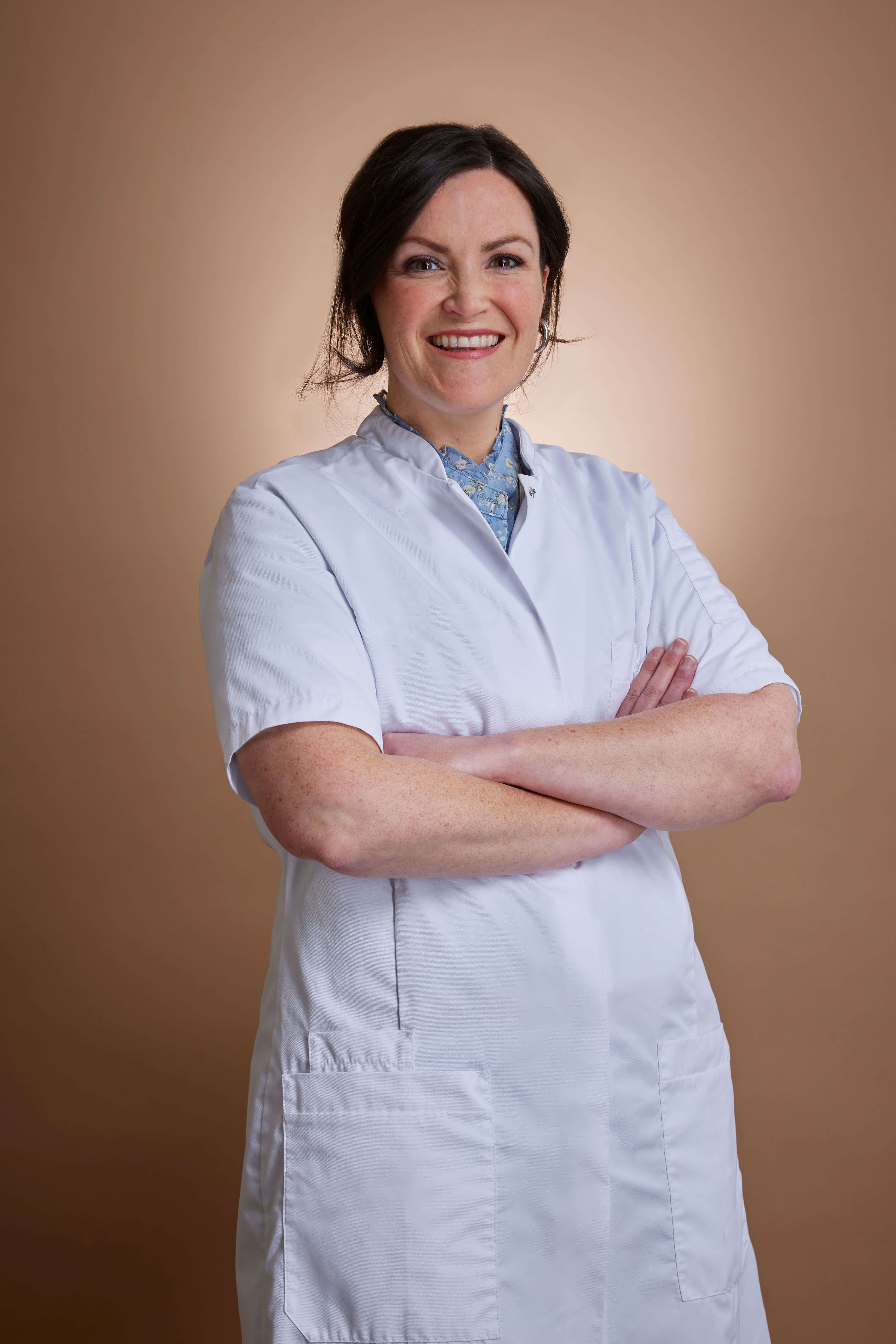 Smiling woman in a white lab coat with crossed arms against a warm brown background.