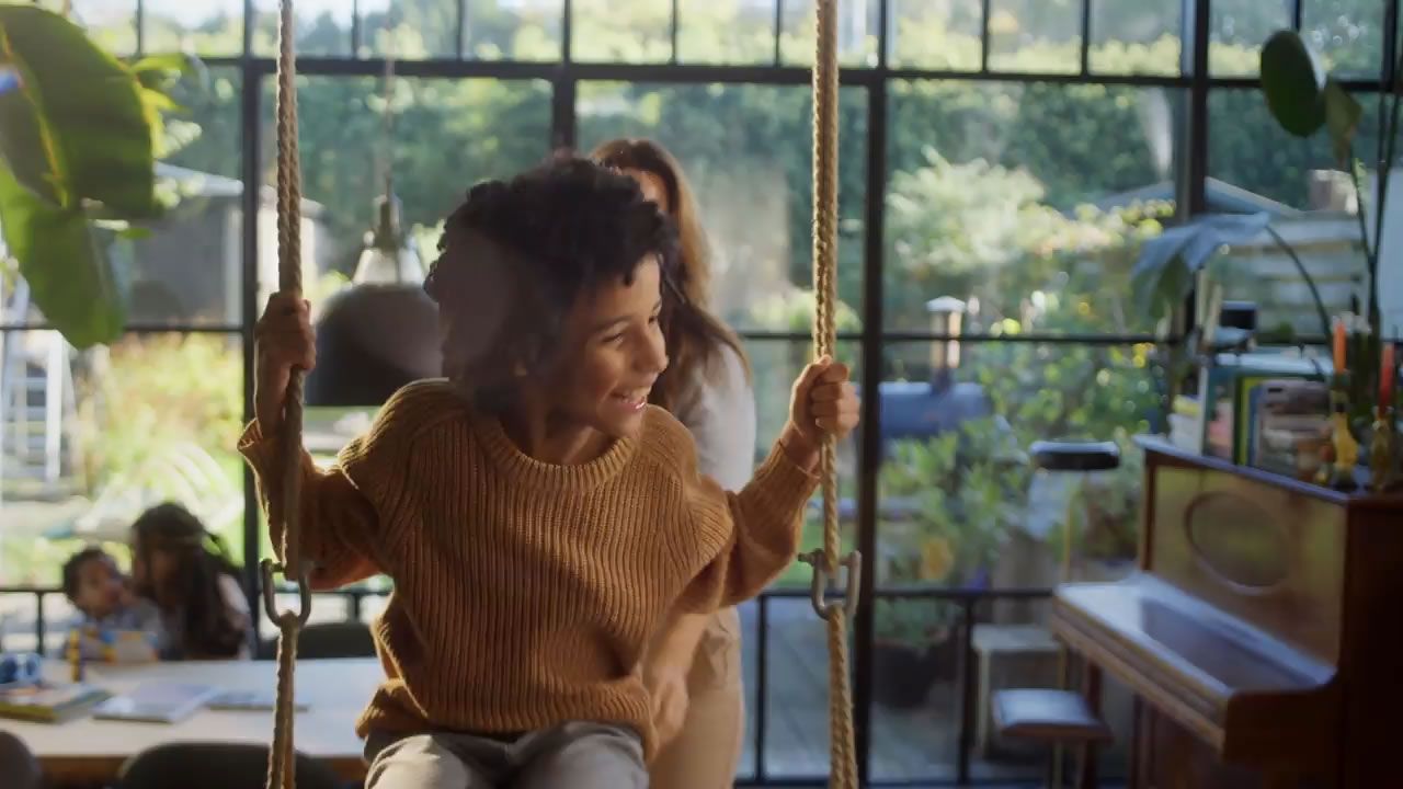 Happy child with curly hair smiles on an indoor swing in a bright, sunlit room.