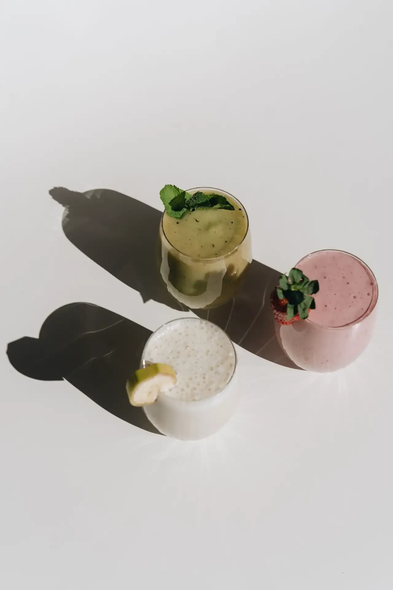 Three small, colorful beverages in minimalist cups with garnishes on a white surface.