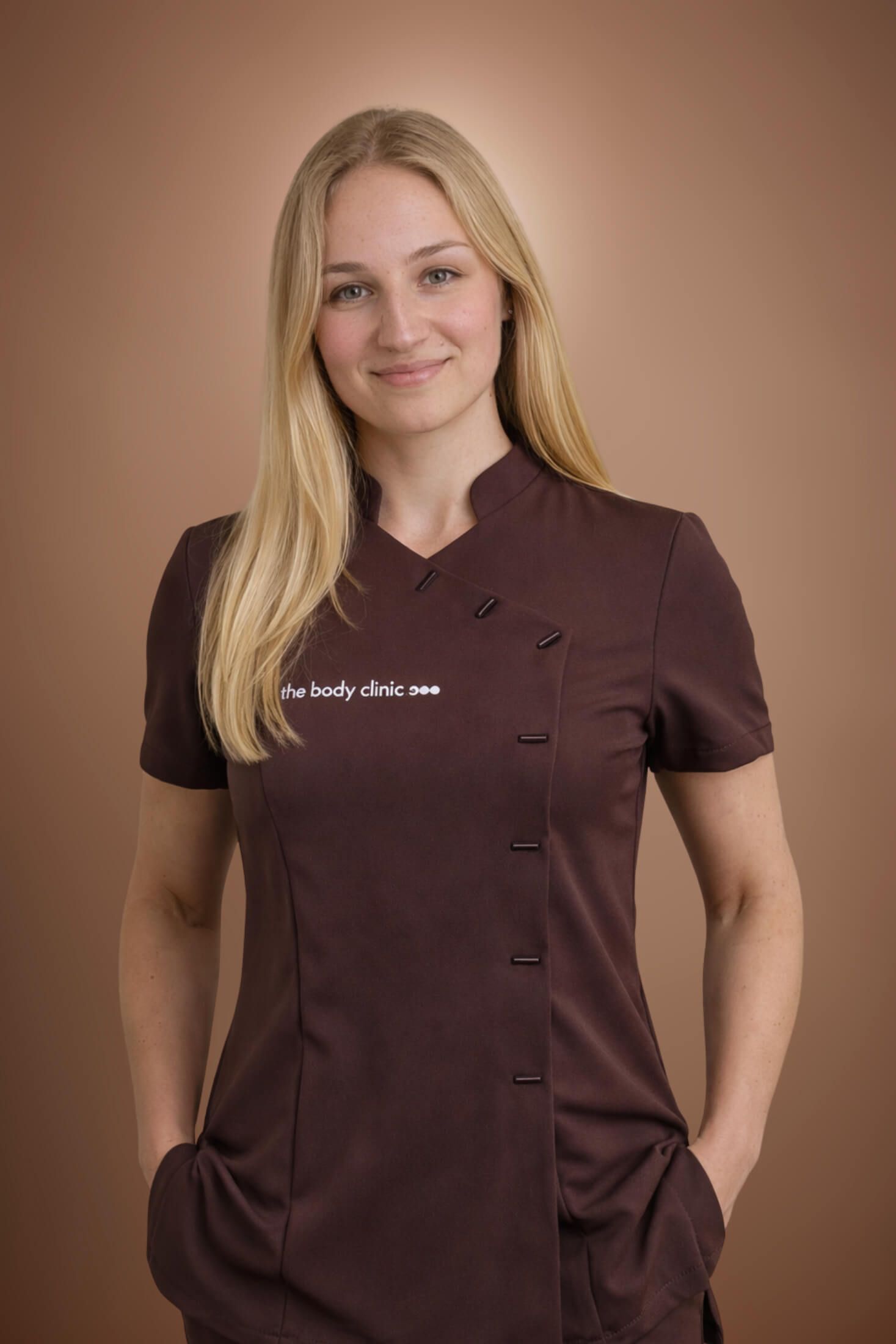 Blonde woman in a dark uniform top, smiling confidently with hands in pockets.