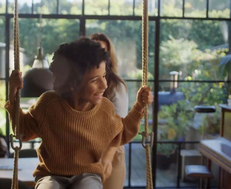 Smiling woman with curly hair on a modern indoor swing, another person visible behind.