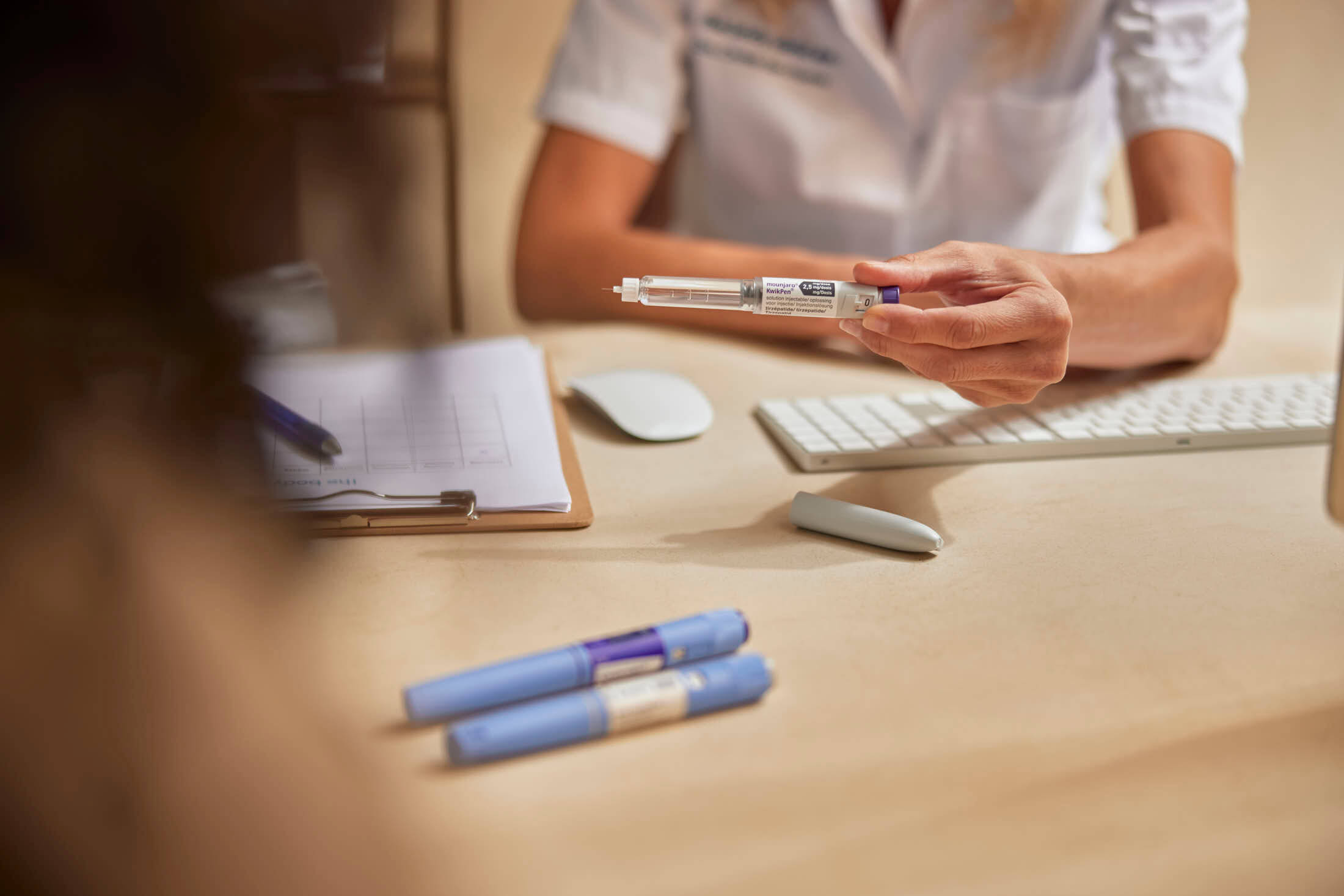 Healthcare professional demonstrates an injection pen to a patient at a desk.