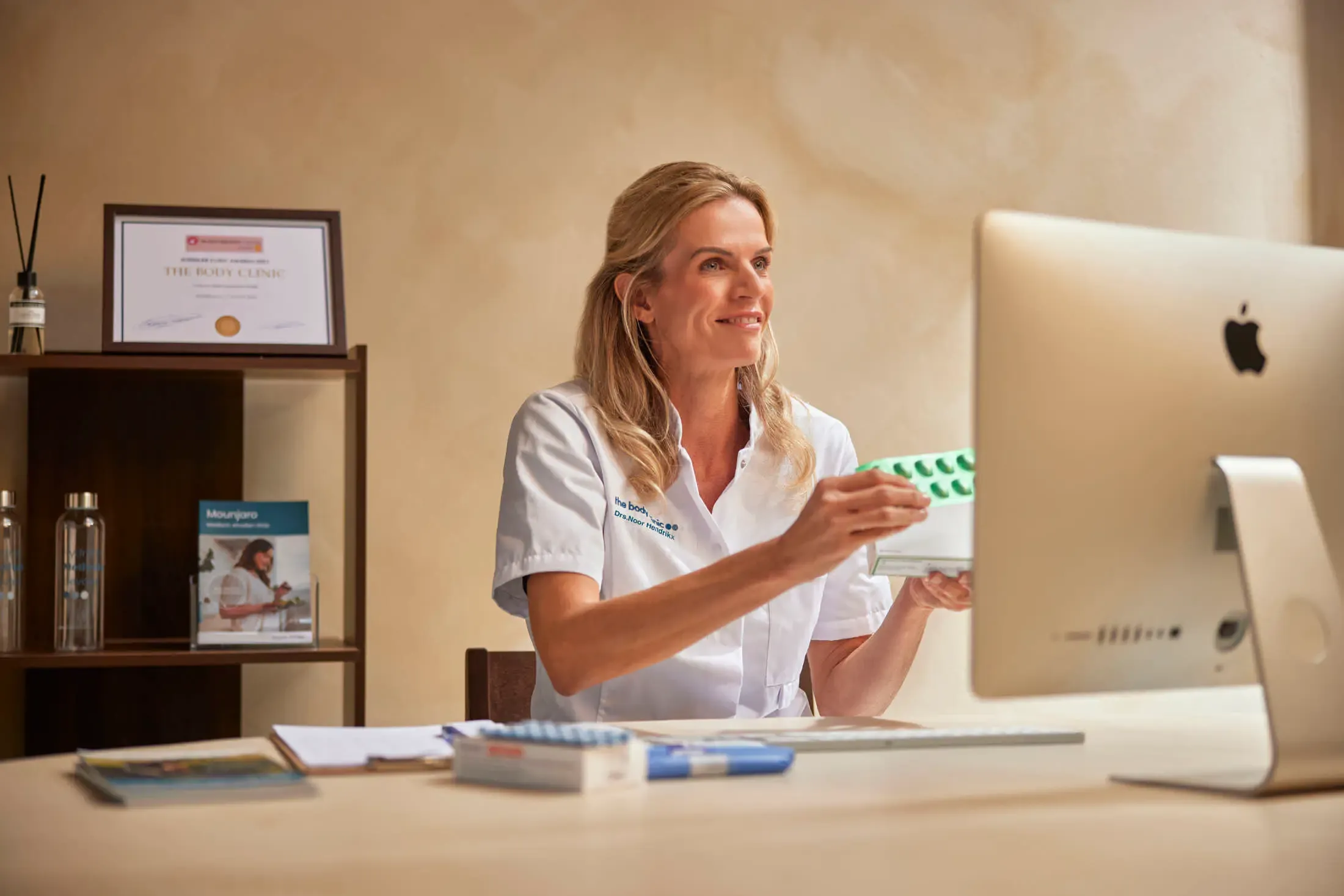 Blonde woman in white uniform holds medication, smiling at her computer screen in an office.