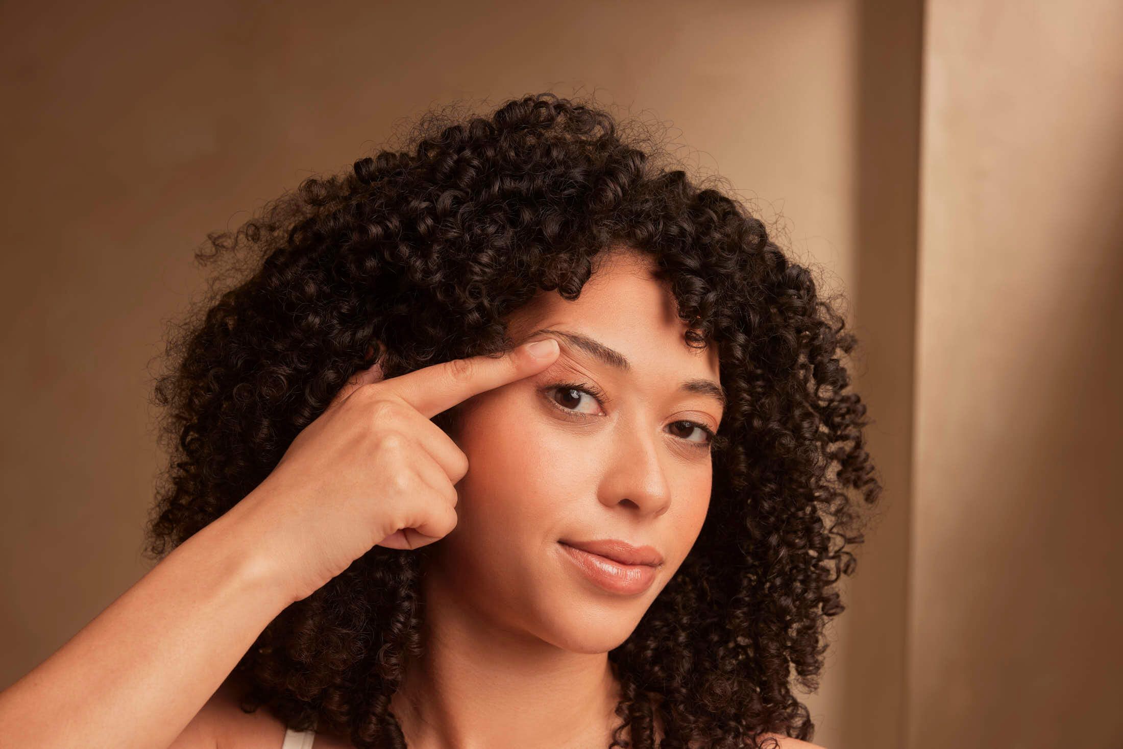 A woman with curly hair points to her eyebrow, demonstrating a beauty routine.