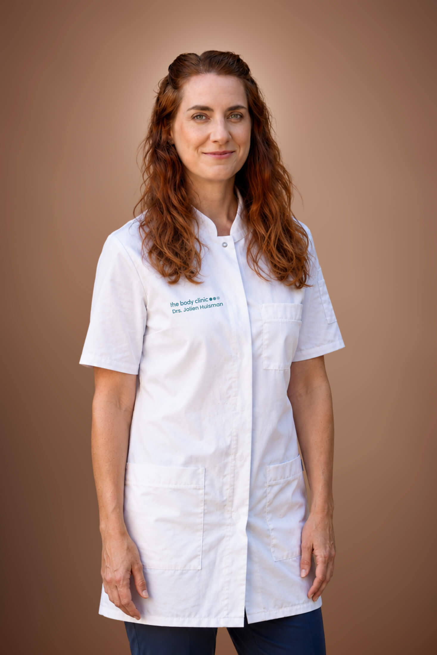 Smiling woman with long brown hair wearing a white medical coat against a brown background.