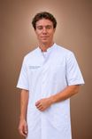 A professional man in a white medical coat stands confidently against a plain brown background.