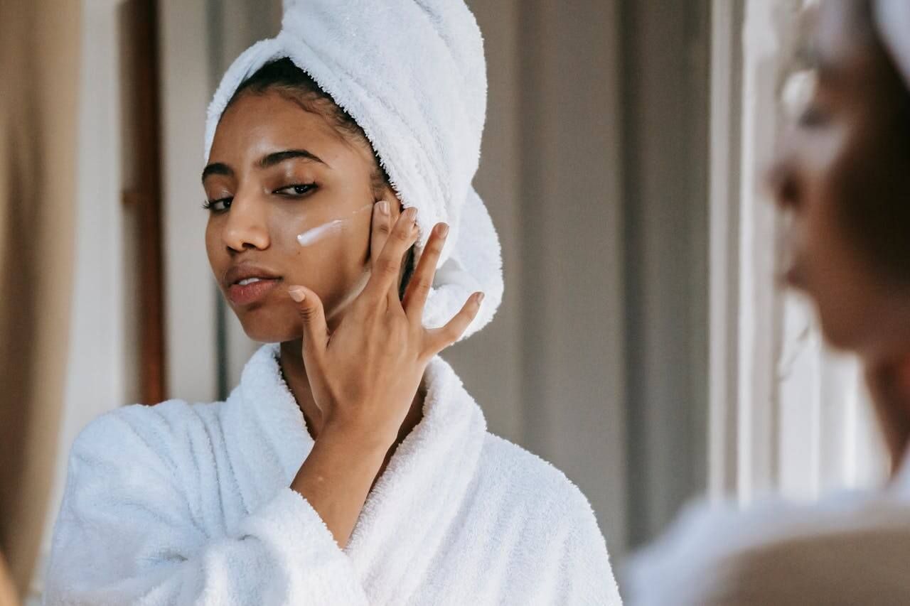 Woman in a white towel and robe applying face cream while looking in a mirror.
