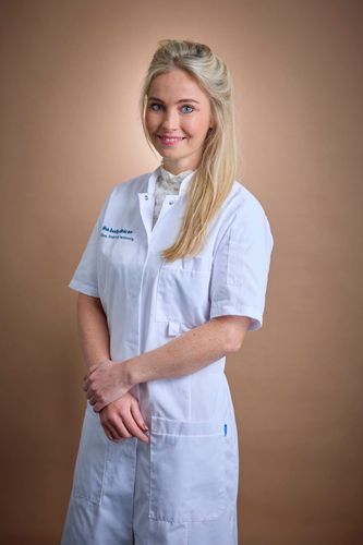 Blonde woman in a white medical coat smiles, hands clasped, looking forward.