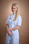 Blonde woman in a white medical coat smiles, hands clasped, looking forward.