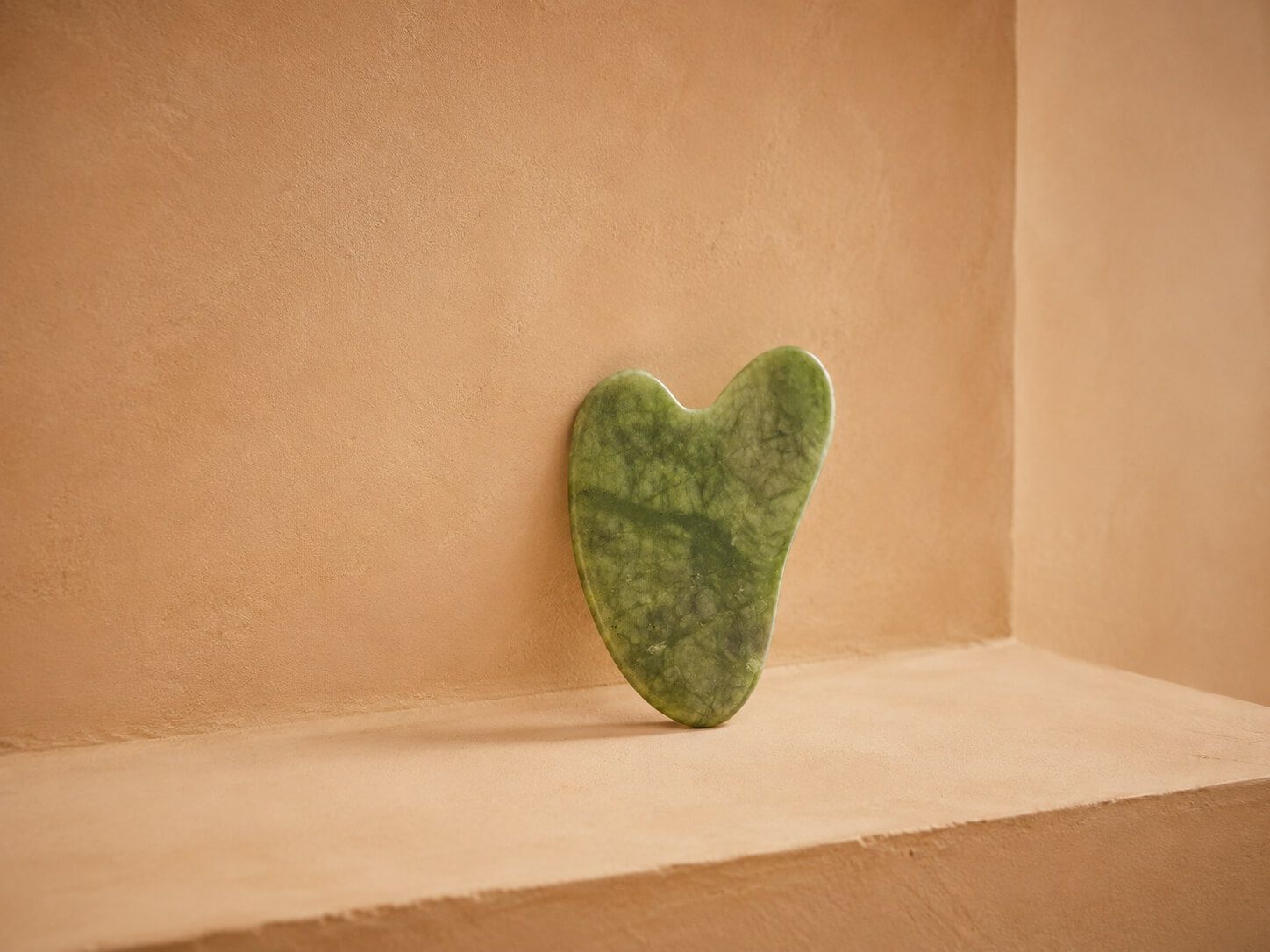 Green jade heart-shaped gua sha tool resting against a warm peach background.