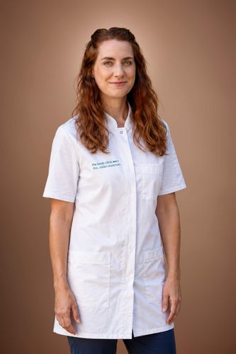 Smiling woman with long brown hair wearing a white medical coat against a brown background.