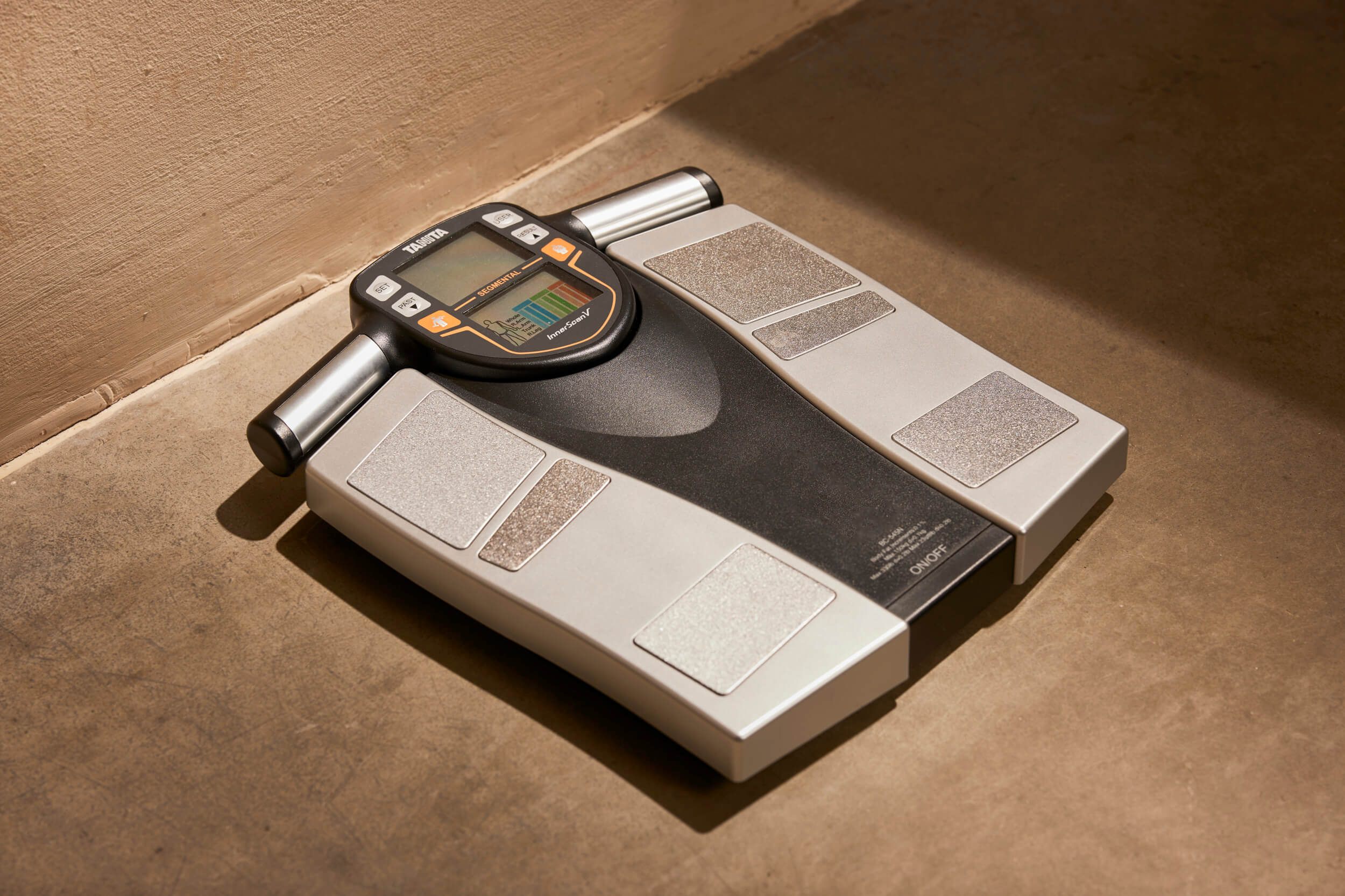 Black and silver digital body composition scale with electrodes on a light brown floor.