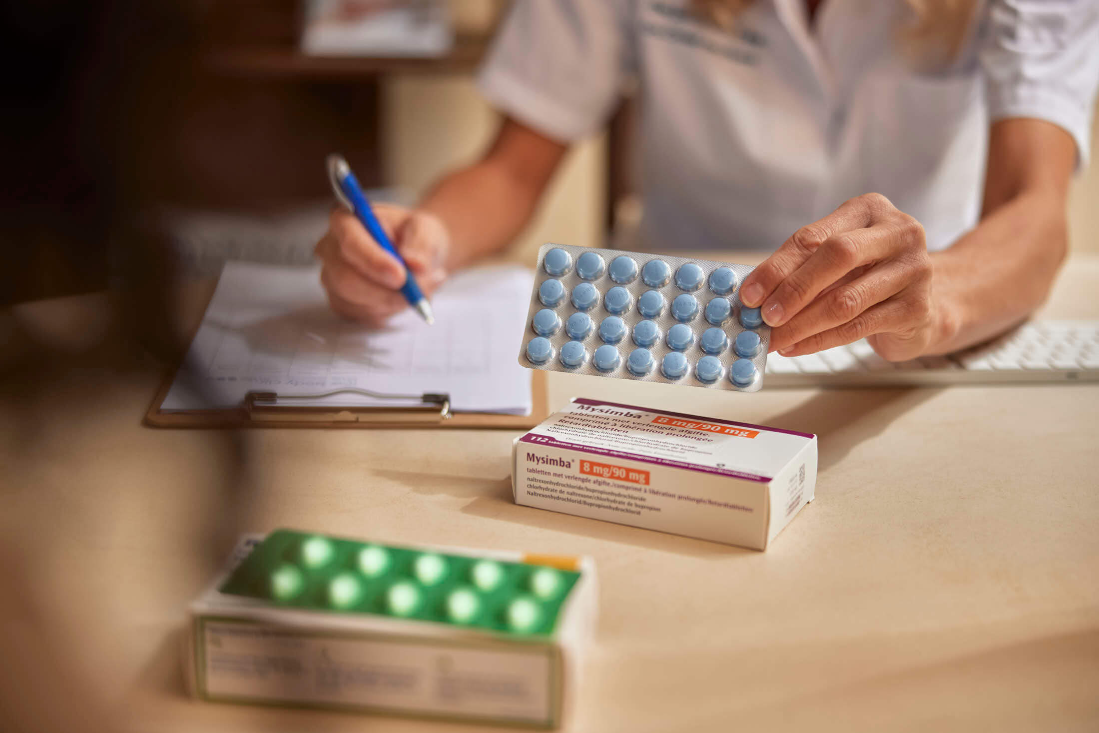 Medical professional holds blue pills while writing on a clipboard with medication boxes.
