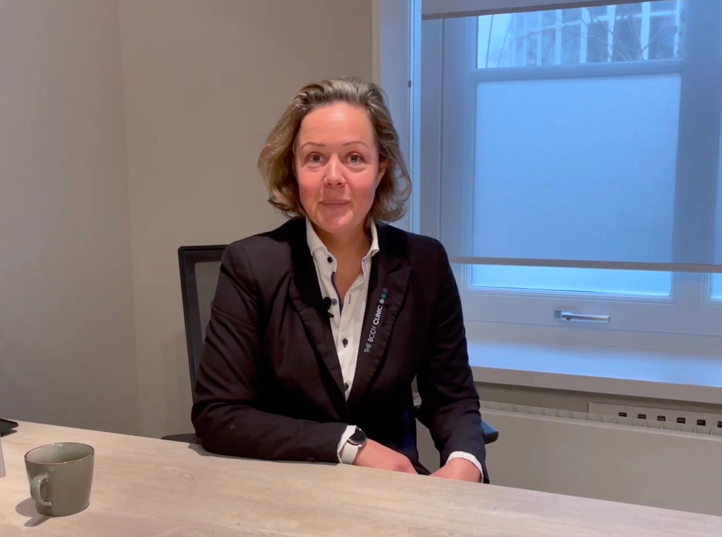 A professional woman in a black blazer sits at a desk, smiling subtly at the camera.