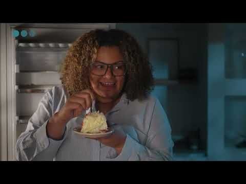 A smiling woman with glasses and curly hair eats a slice of cake from an open refrigerator.