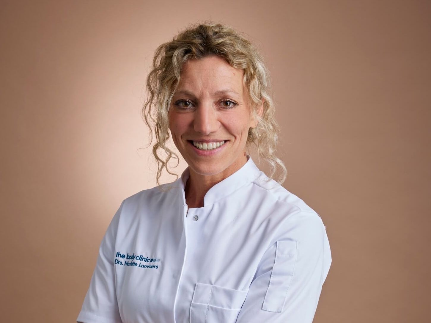 Smiling female doctor with curly blonde hair wearing a white medical coat.