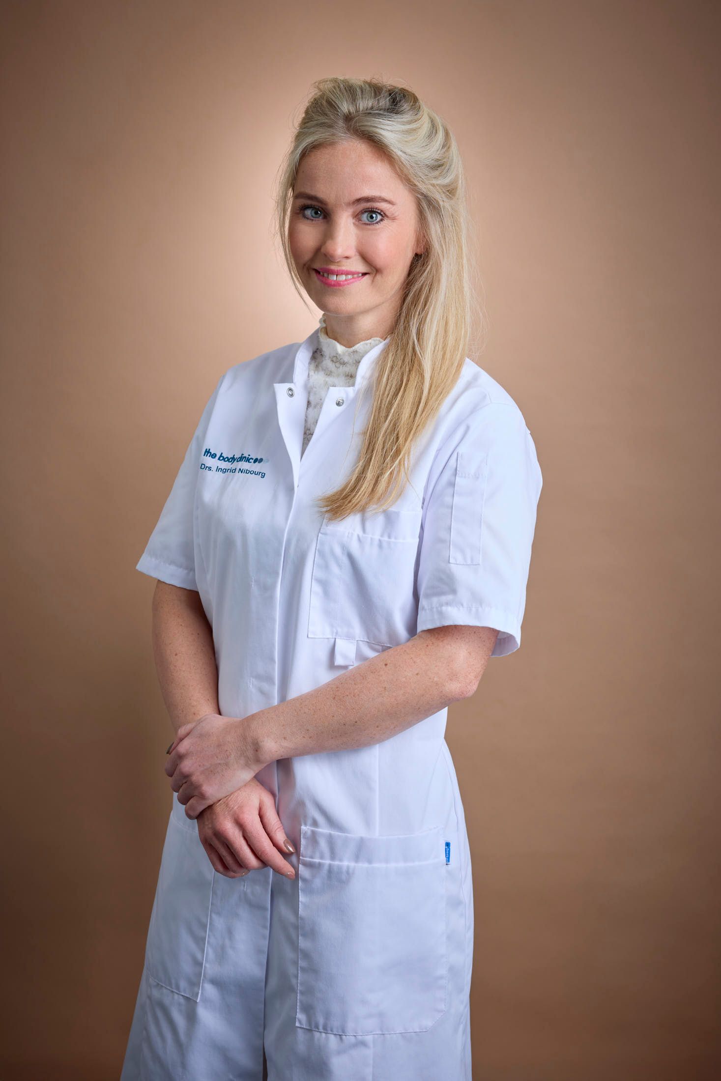 Blonde woman in a white medical coat smiles, hands clasped, looking forward.