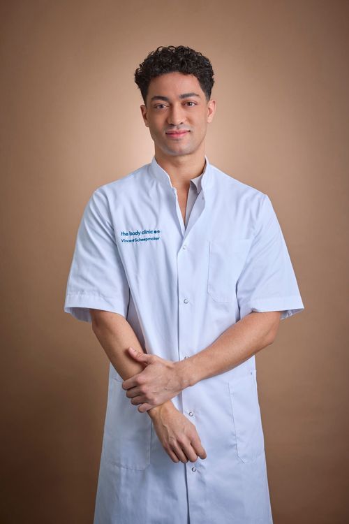 Young man in light blue medical coat stands confidently, looking at the camera.