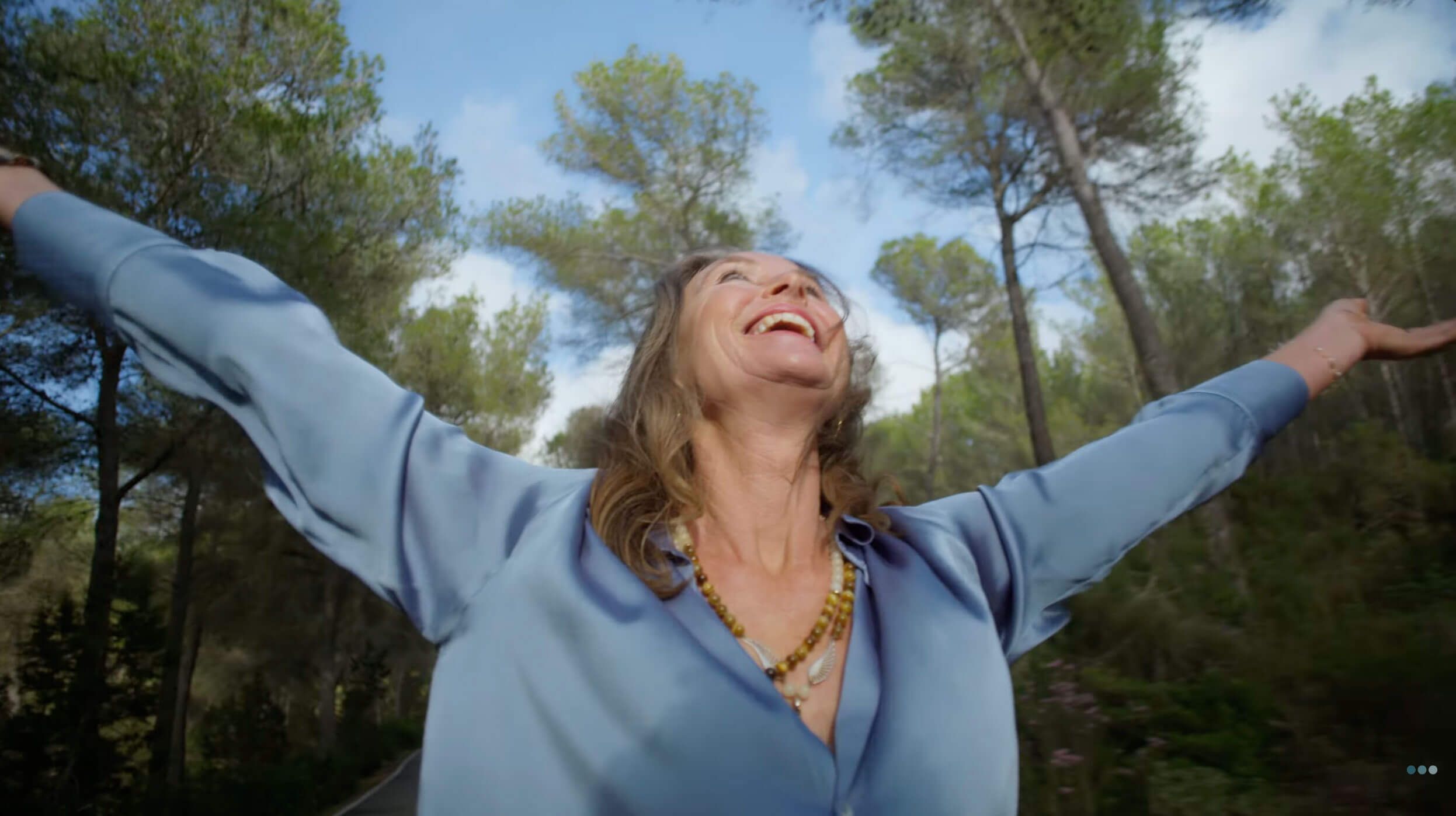 A joyful woman with outstretched arms smiles up at the sky amidst green trees.