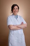 Smiling woman in a white lab coat with crossed arms against a warm brown background.